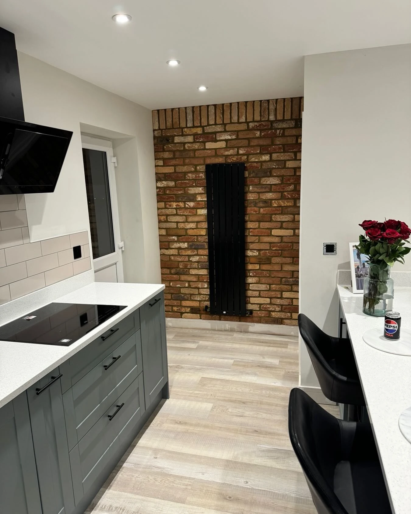 Modern kitchen with gray cabinets, white counters, wood floor, brick wall, black radiator, and black chairs.