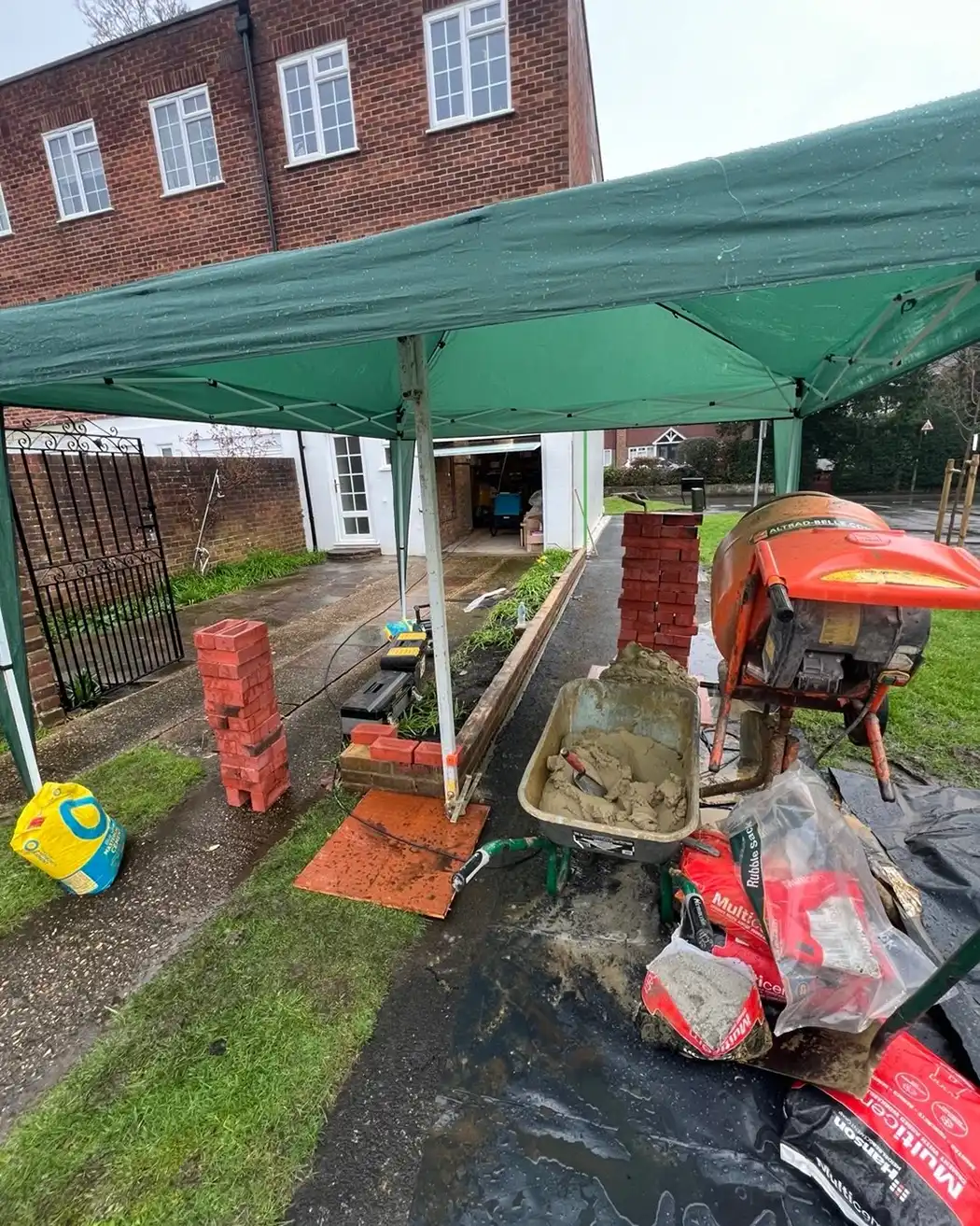 Outdoor construction setup with bricks, cement mixer, wheelbarrow with mortar under green canopy.