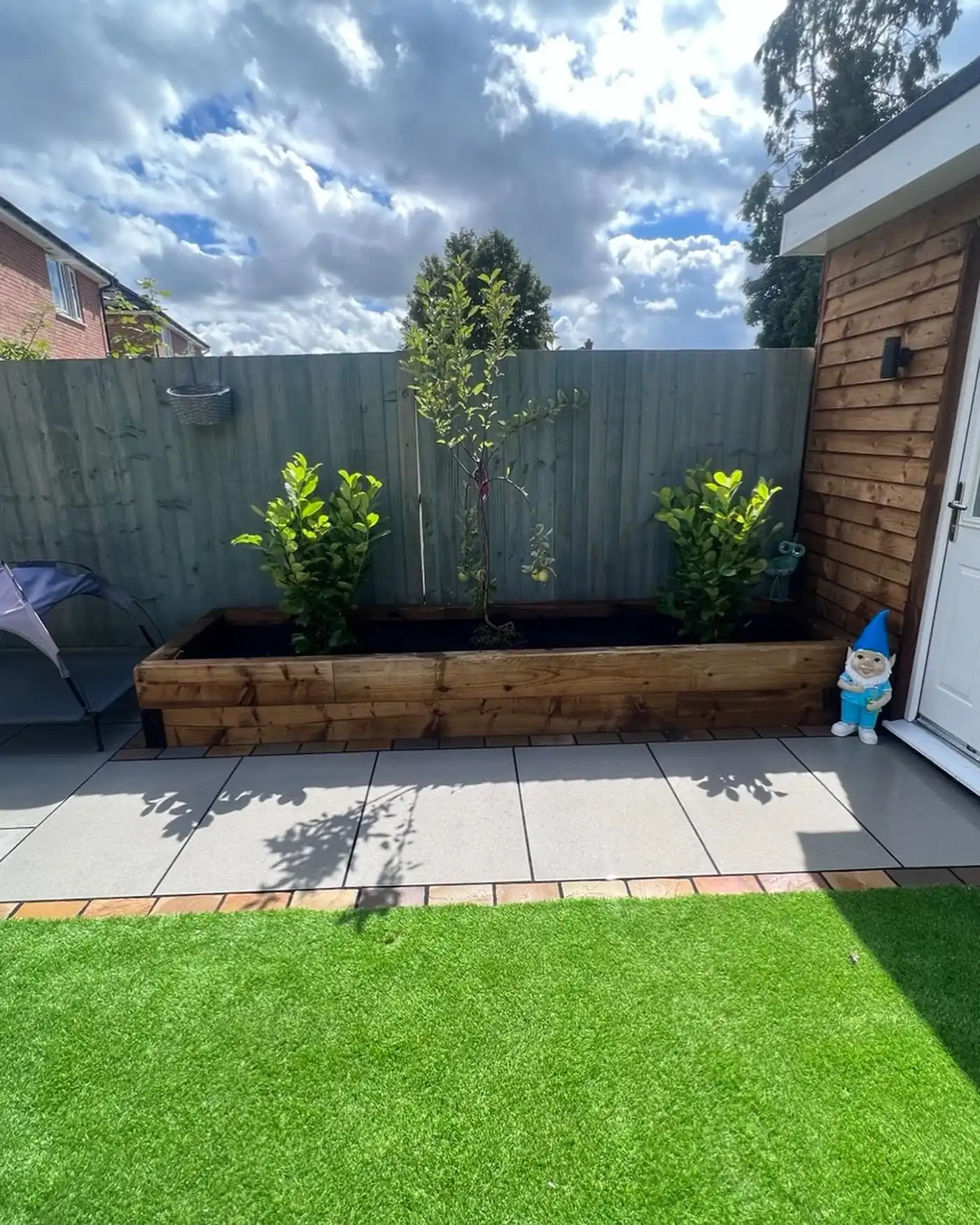 Backyard with green artificial grass, paved area, raised wooden planter with lemon tree and shrubs, blue garden gnome.
