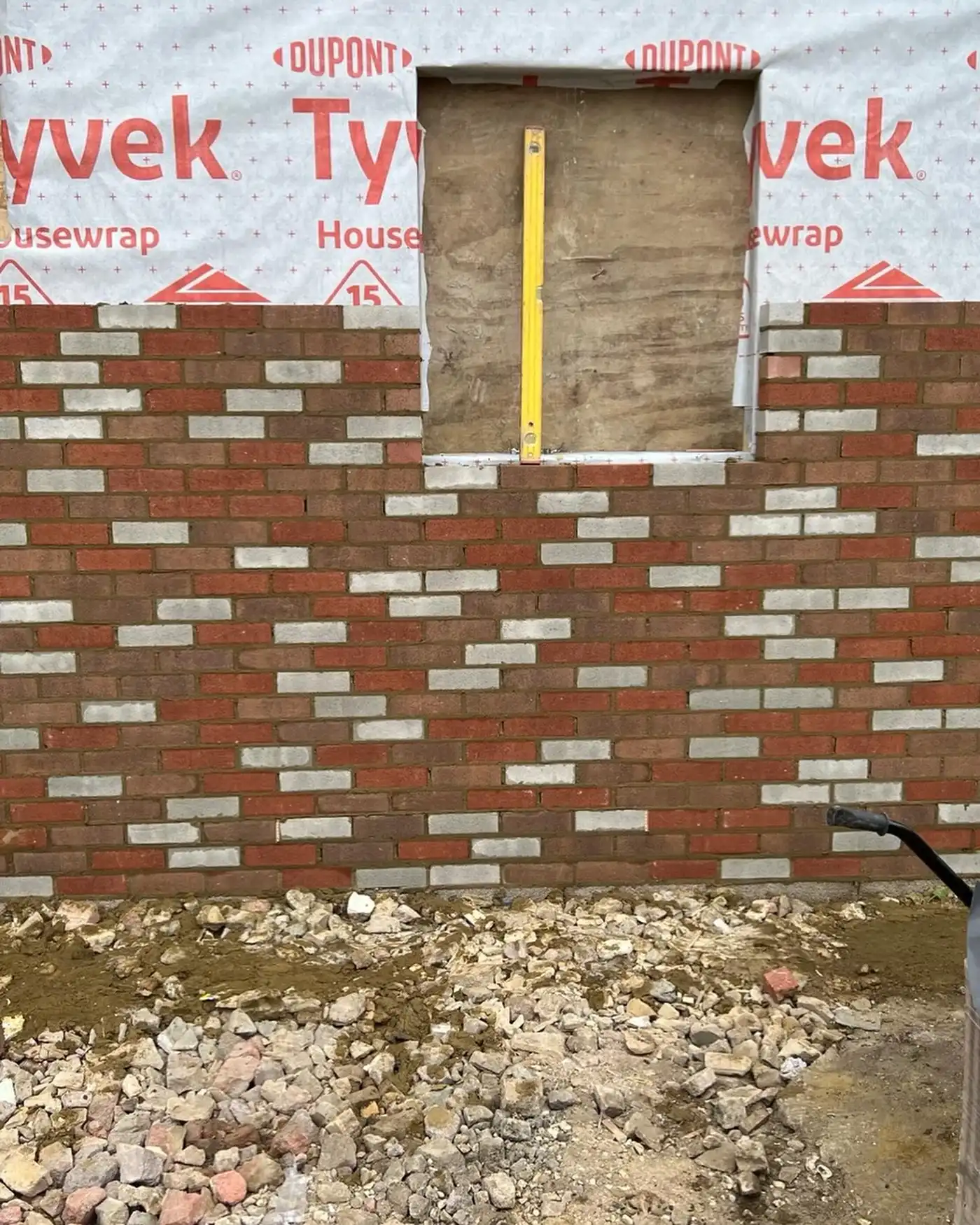 Wall under construction with red and gray bricks, window opening, and rocky ground in front.