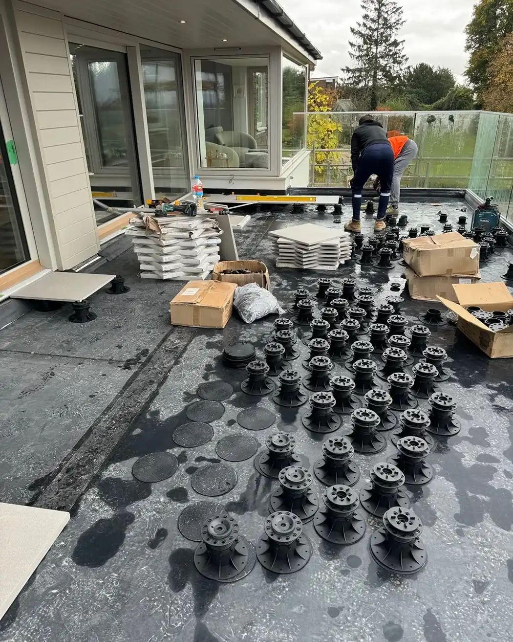 Two workers installing black pedestal supports for raised flooring on a balcony with stacked tiles nearby.