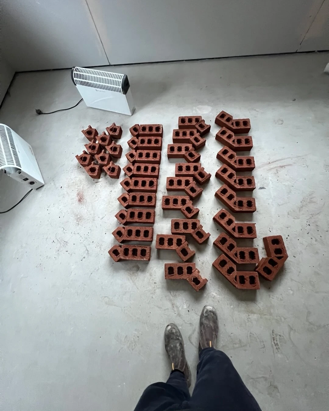 Organized stacks of small red bricks on a concrete floor with two white heaters nearby.