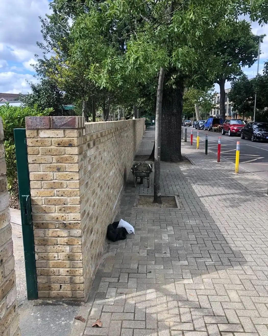 Paved sidewalk beside a brick wall with trees, a wheelbarrow with soil, and parked cars on the street.