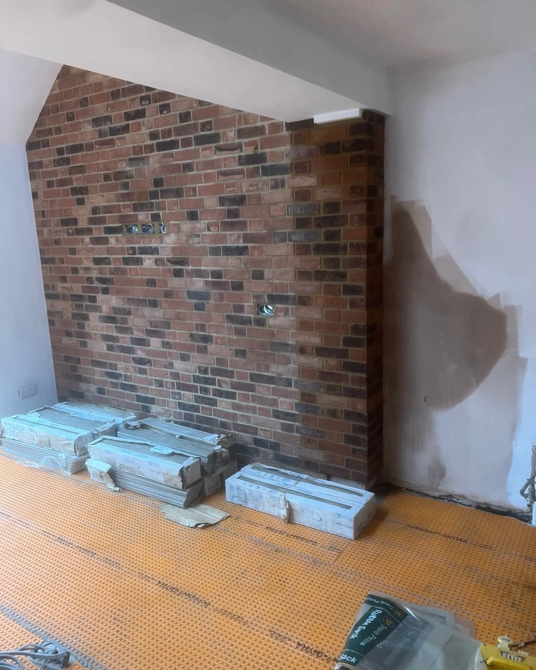 Room under renovation with exposed brick wall, white walls, orange floor protection, and stacked construction materials.