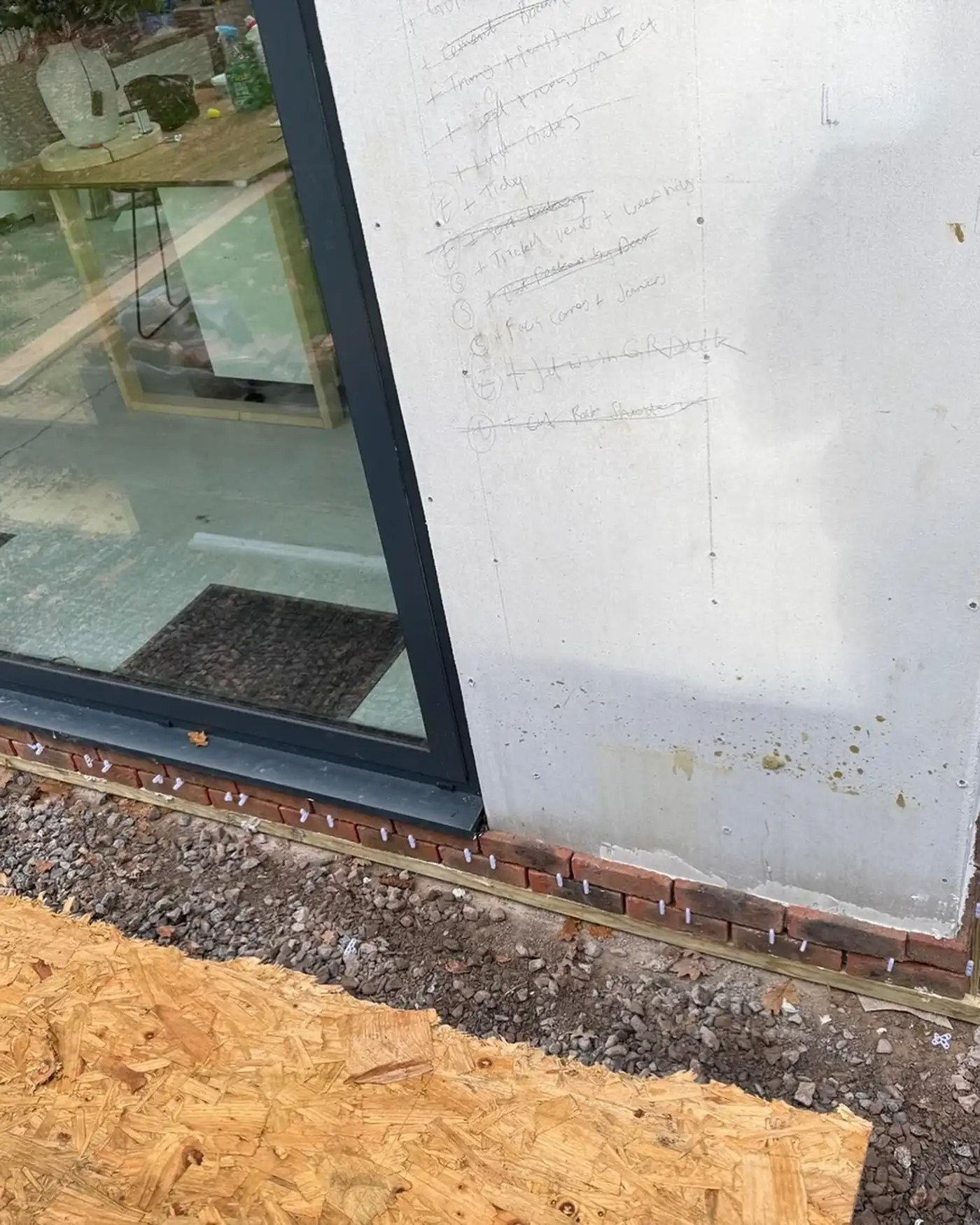 Construction site showing brick foundation laid beside a glass door with handwritten notes on wall.