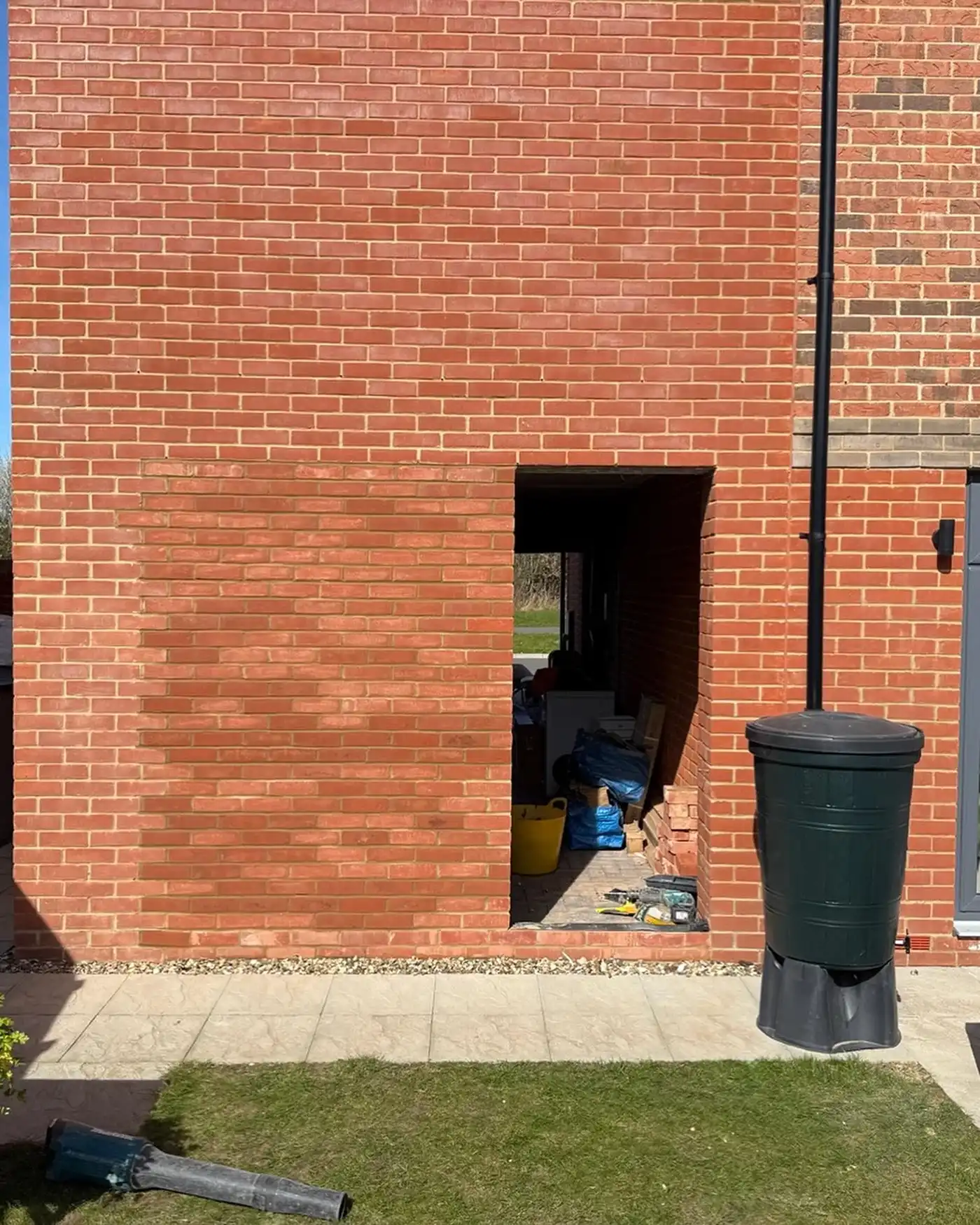 Brick wall with a rectangular opening showing construction materials and a black rain barrel nearby.