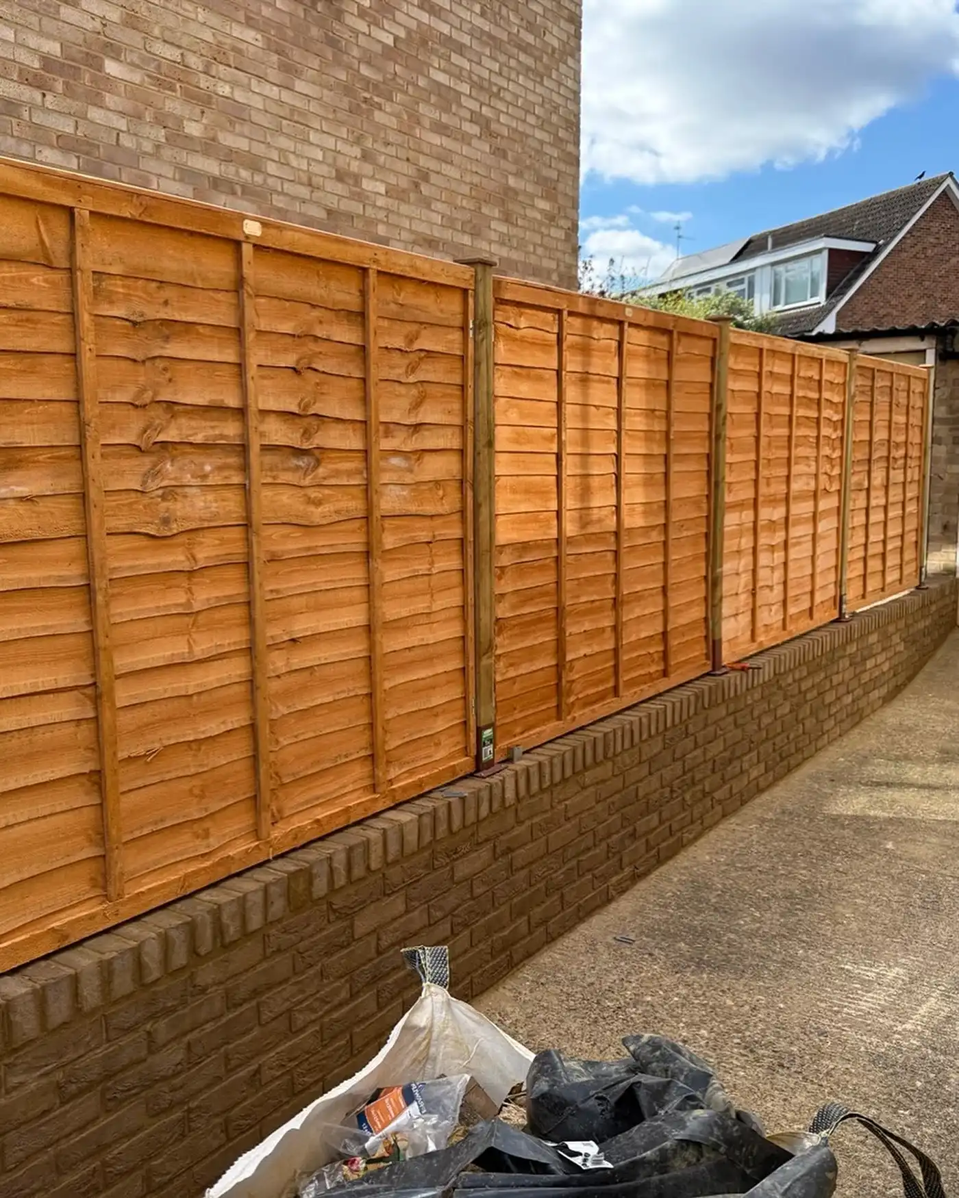 New wooden fence installed on top of a low brick wall along a paved driveway with buildings in the background.