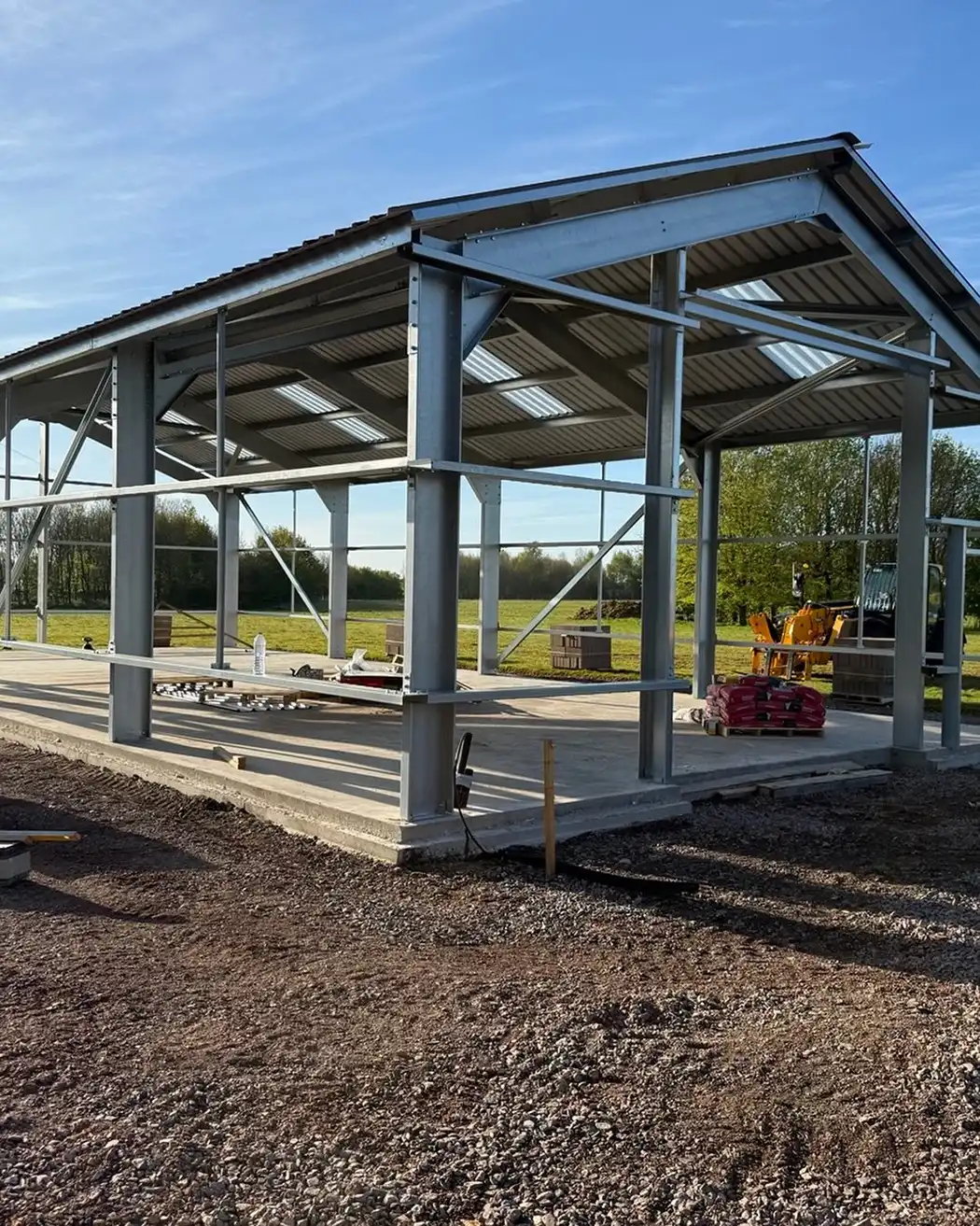 Steel frame structure under construction on concrete slab in an open outdoor area with green grass and trees.