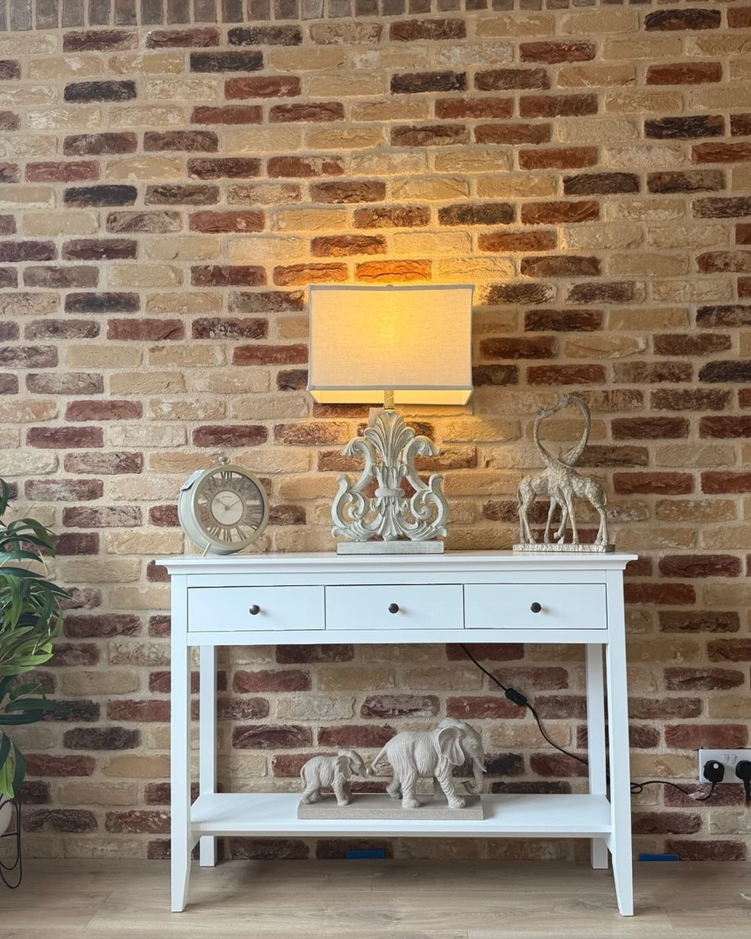 White console table with a lamp, clock, giraffe sculpture, and elephant figurines against a brick wall.