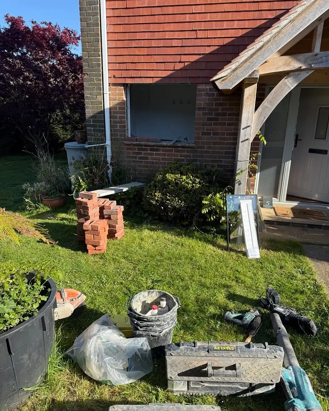 Construction tools and bricks on grass in front of a house with a window opening and wooden porch.