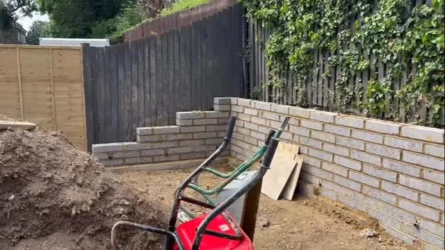 Partially built brick wall in backyard with a wheelbarrow and a pile of dirt nearby.