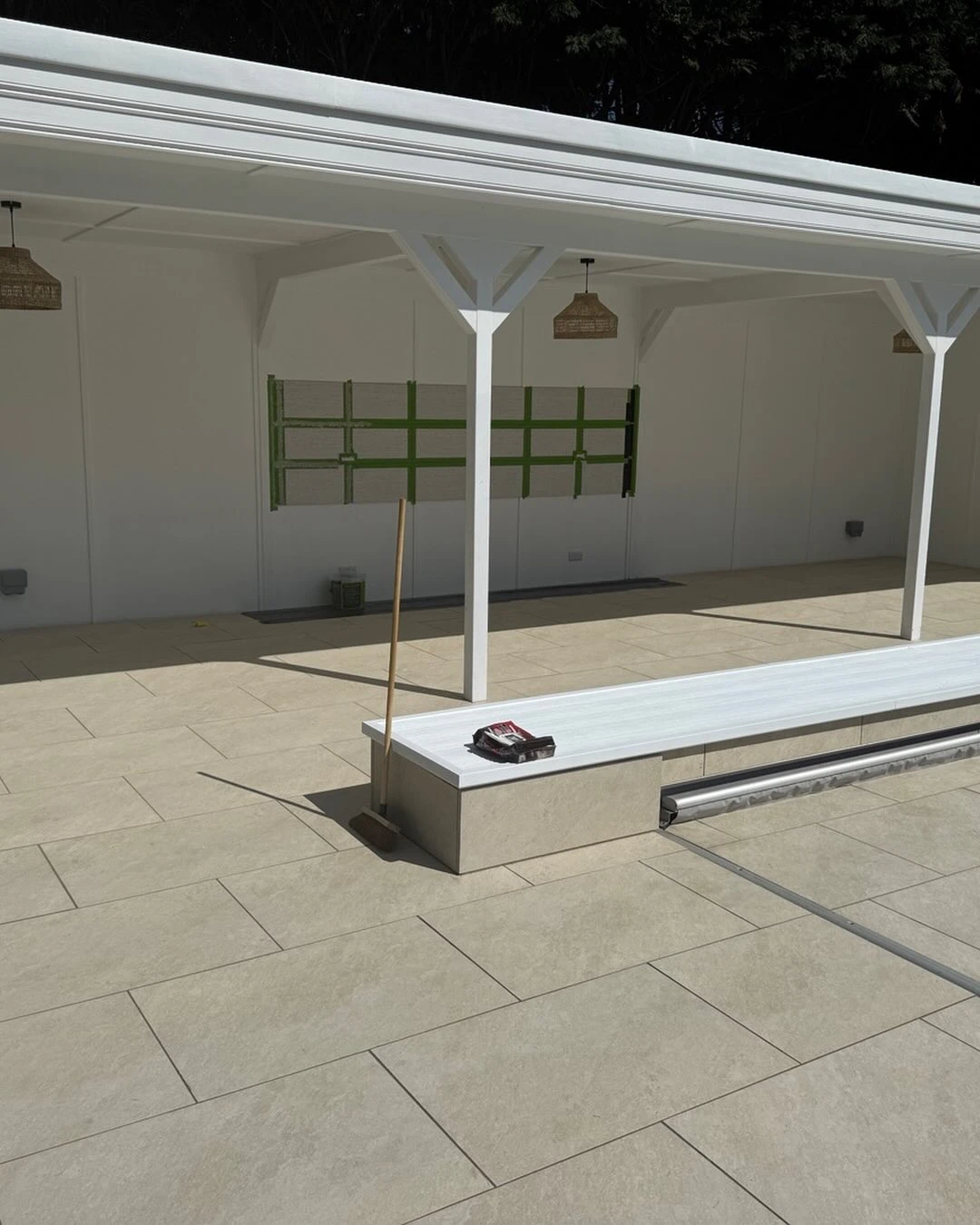 Outdoor tiled patio with white pergola, broom leaning on bench, and garden tools on the bench.