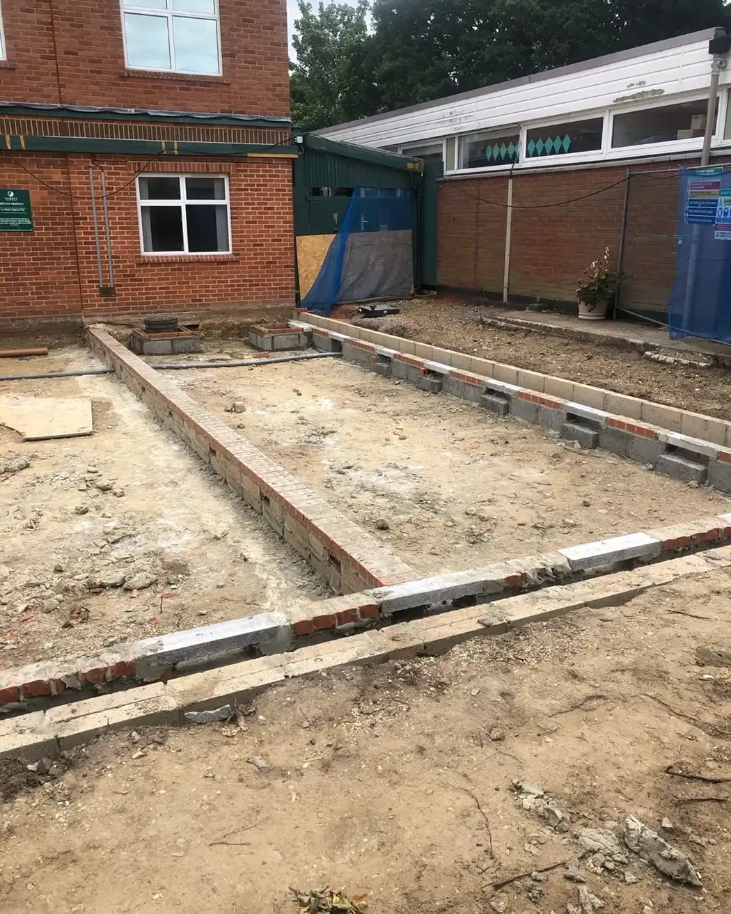 Construction site showing foundation walls and cleared ground next to brick buildings.